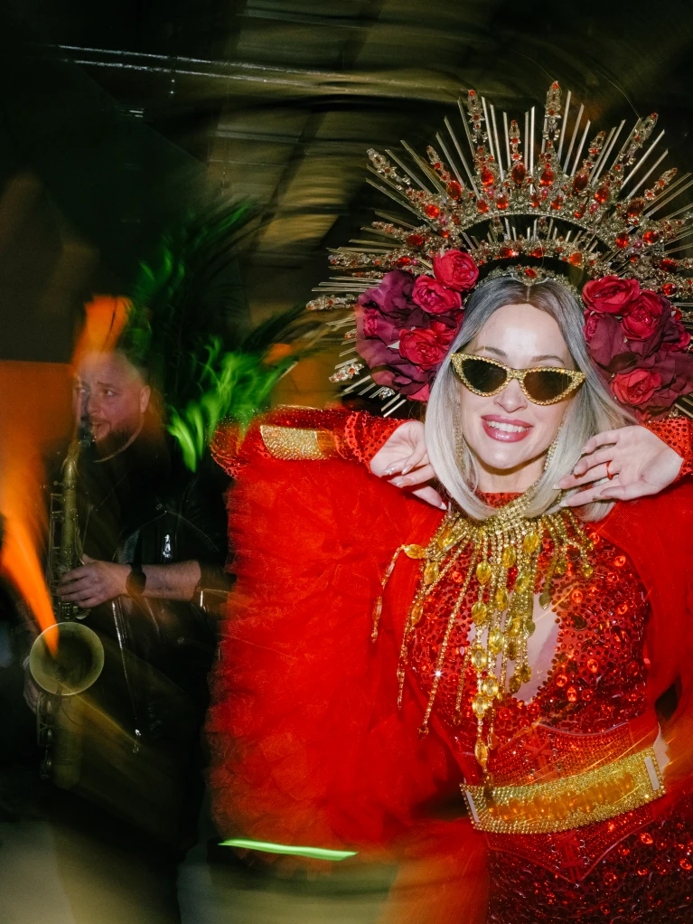 Frau in rotem Karnevalskostüm mit Kopfschmuck aus Rosen und Sonnenbrille, posiert lächelnd, unscharfer Hintergrund mit Saxophonspieler