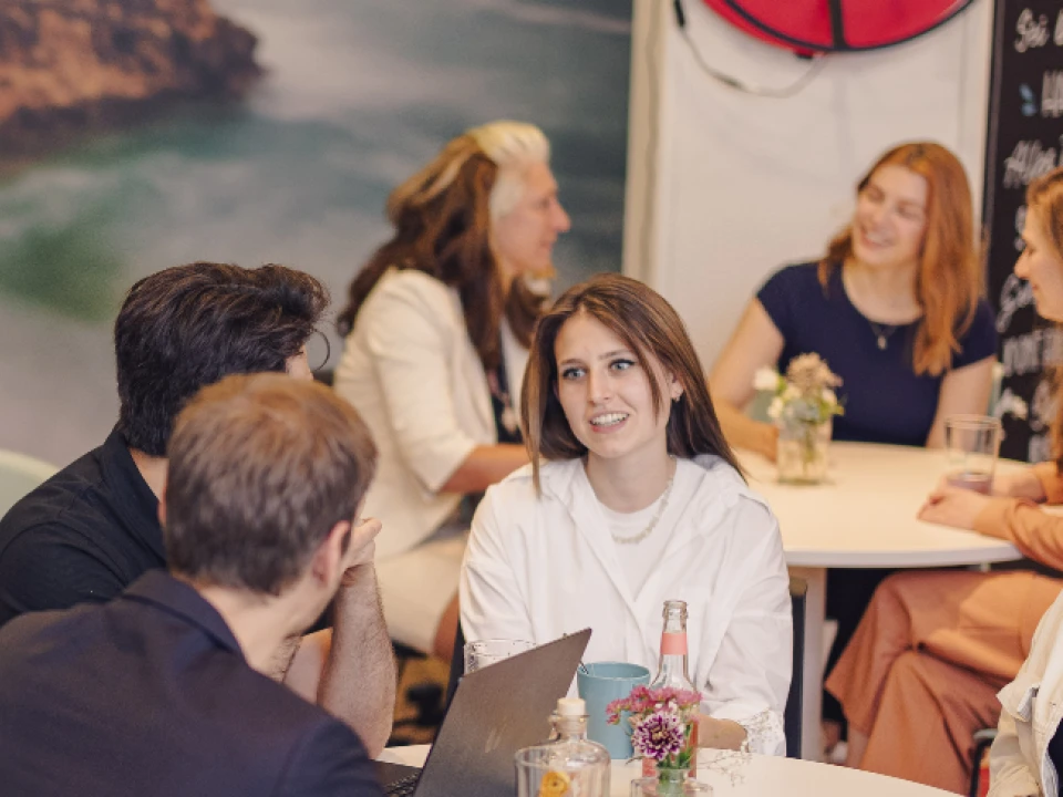 Gruppe von Menschen in einem informellen Bürotreffen, sitzend an Tischen, unterhaltend, mit einer großen Wandgrafik im Hintergrund.