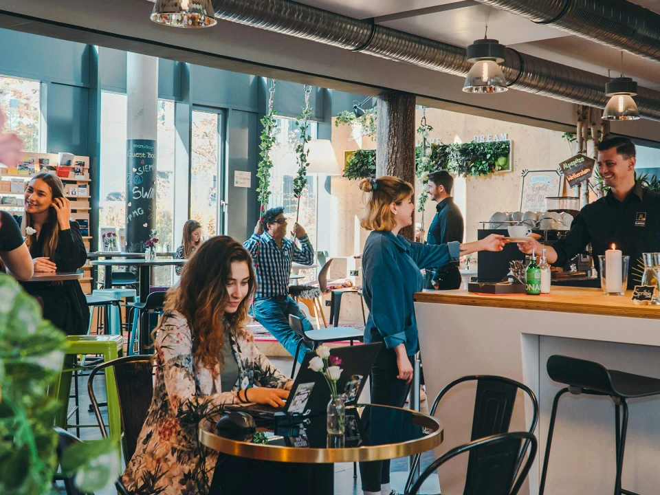 Modernes Café mit urbanem Design, in dem Menschen Kaffee genießen, arbeiten und sich unterhalten. Ein Barista serviert einer Kundin einen Kaffee an der Theke.