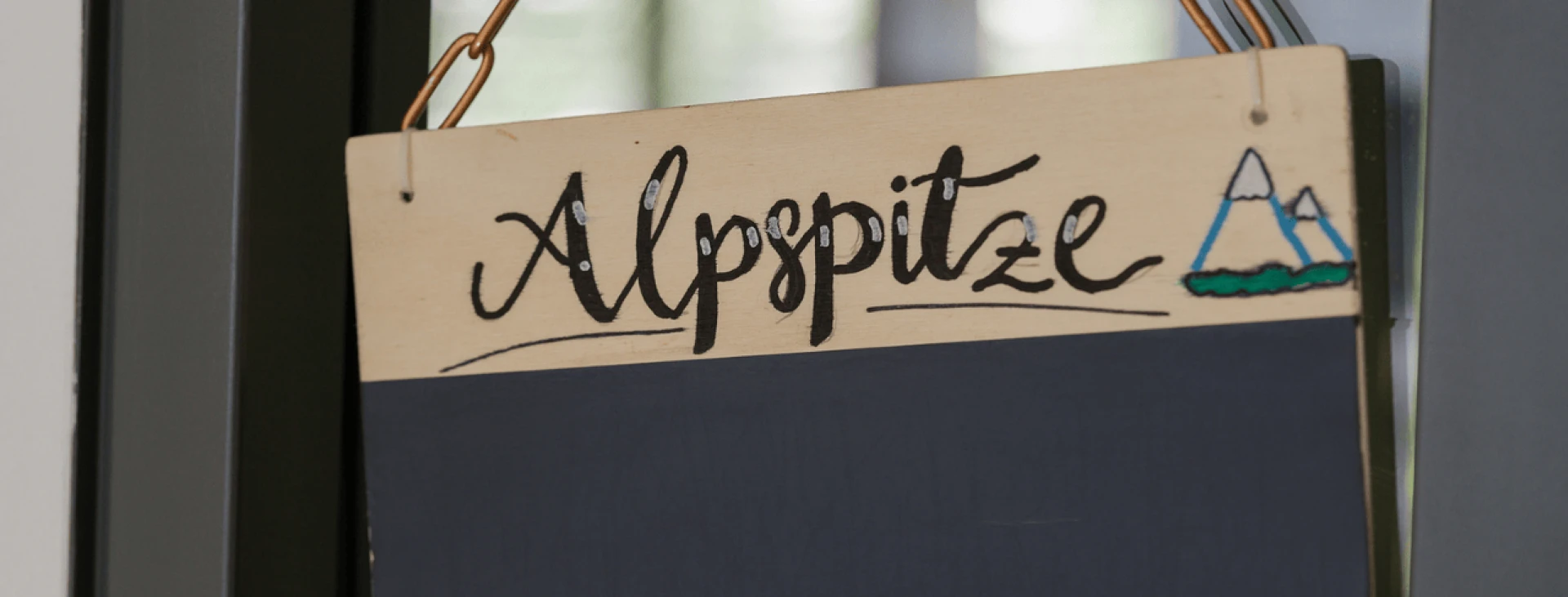 Ein hölzernes Schild mit der Aufschrift 'Alpspitze' und einer gemalten Bergsilhouette neben einem Zelt.