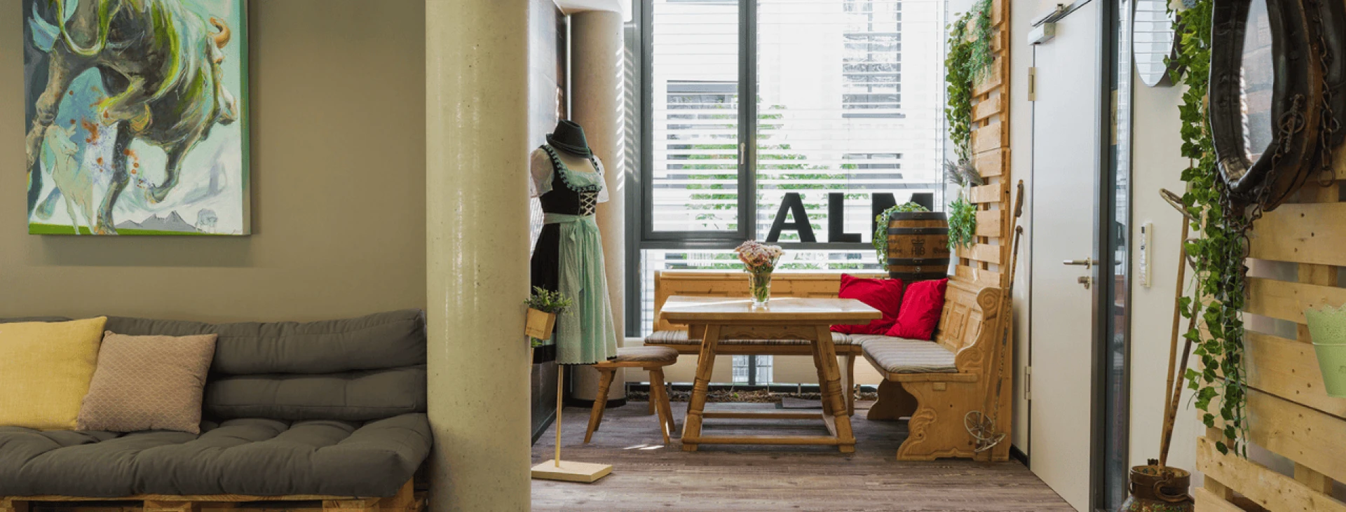 Bohemisch eingerichtetes Wohnzimmer mit grauem Sofa, Holztisch, roter Bank und zahlreichen Pflanzen. An der Wand hängen Bilder und ein Spiegel.