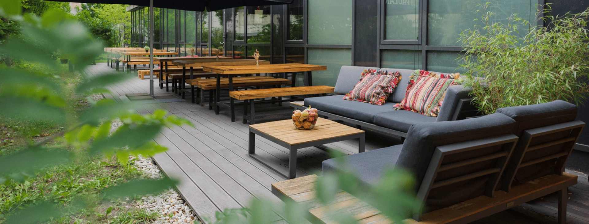 Holzterrasse mit mehreren Sitzbereichen, darunter Holztische und Bänke sowie eine Loungeecke mit grauen Sofas und bunten Kissen, umgeben von Grün