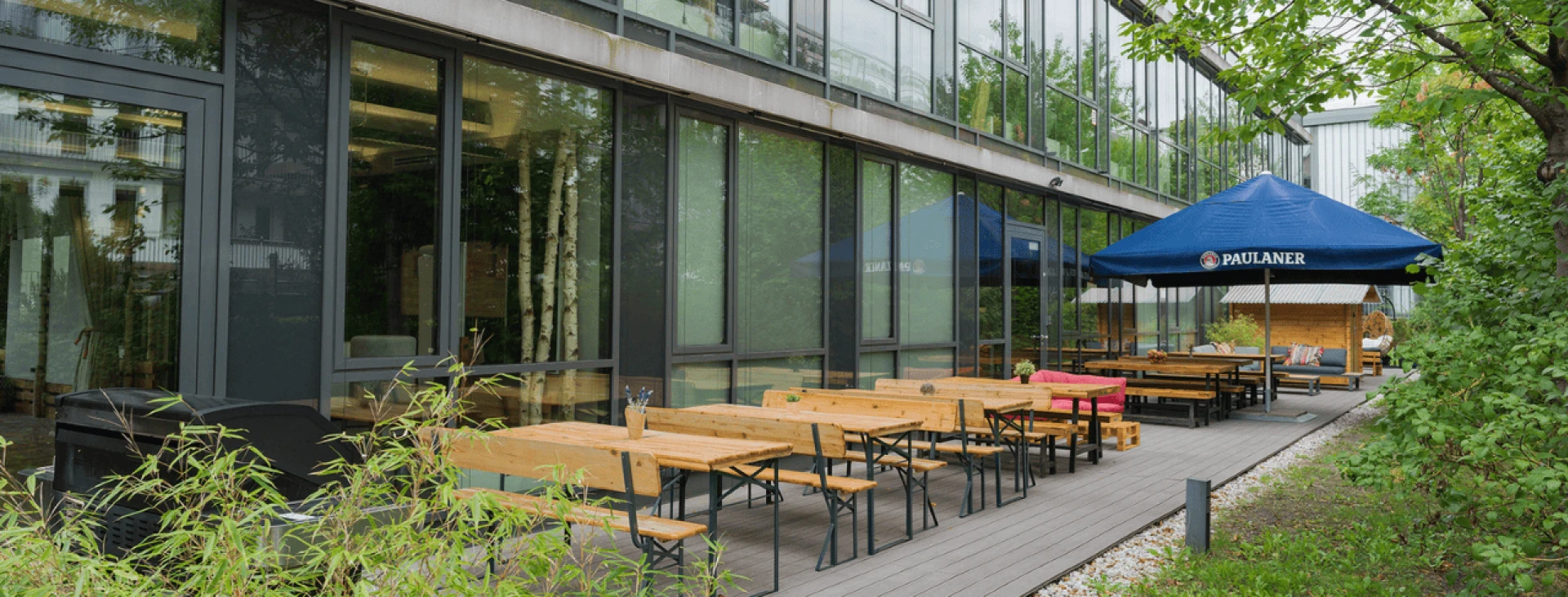 Außenbereich eines modernen Gebäudes mit einer Terrasse, die mit Holztischen und -bänken bestückt ist, unter einem blauen Sonnenschirm