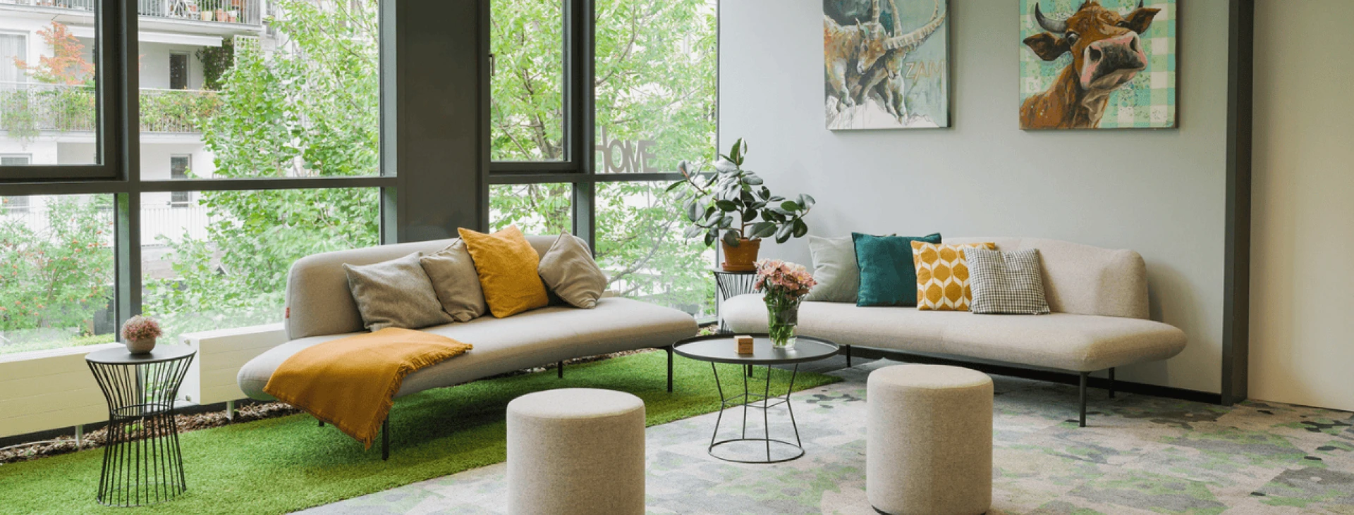 Modern eingerichtetes Wohnzimmer mit zwei Sofas in Beige, dekoriert mit gelben und gemusterten Kissen, runden Hockern, einem grünen Teppich und Pflanzen neben großen Fenstern.