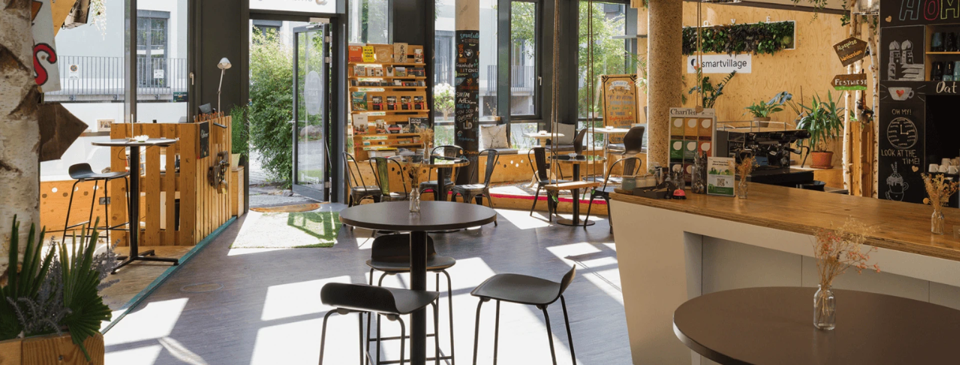 Innenansicht eines modernen Cafés mit verschiedenen Sitzbereichen, offener Tür und einer Theke mit Getränken und Snacks.