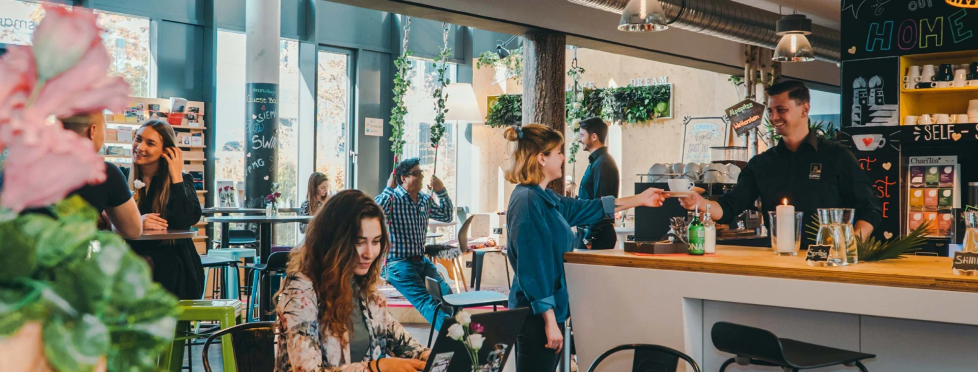 Lebhaftes Café mit mehreren Gästen, die an Tischen sitzen und mit Barpersonal sprechen. Moderne Einrichtung mit Pflanzen und Dekor.