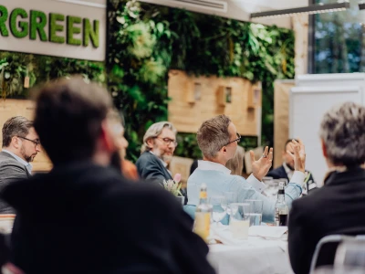 Personen sitzen an einem langen Tisch bei einer Konferenz in einem Raum mit viel Grün und einem Schild 'EVERGREEN'. Einige Personen gestikulieren während der Diskussion.