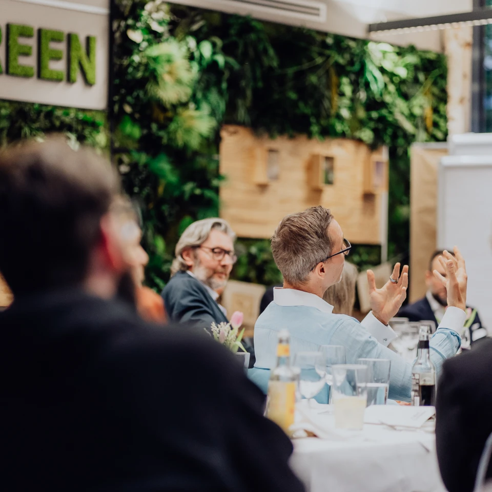 Personen sitzen an einem langen Tisch bei einer Konferenz in einem Raum mit viel Grün und einem Schild 'EVERGREEN'. Einige Personen gestikulieren während der Diskussion.