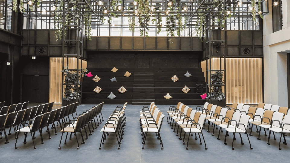Modern event hall with rows of black and white chairs facing a stage with suspended art installations and green plant decorations in the upper area.