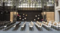 Modern event hall with rows of black and white chairs facing a stage with suspended art installations and green plant decorations in the upper area.