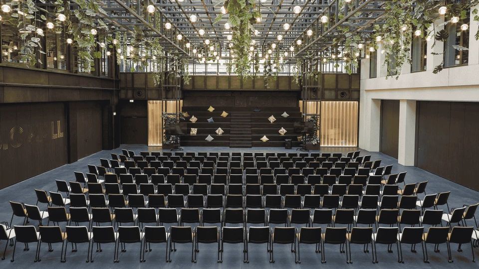 Modern performance hall with a semicircular seating arrangement of black chairs, stage at the front, covered by a glass roof with hanging plants and lights.