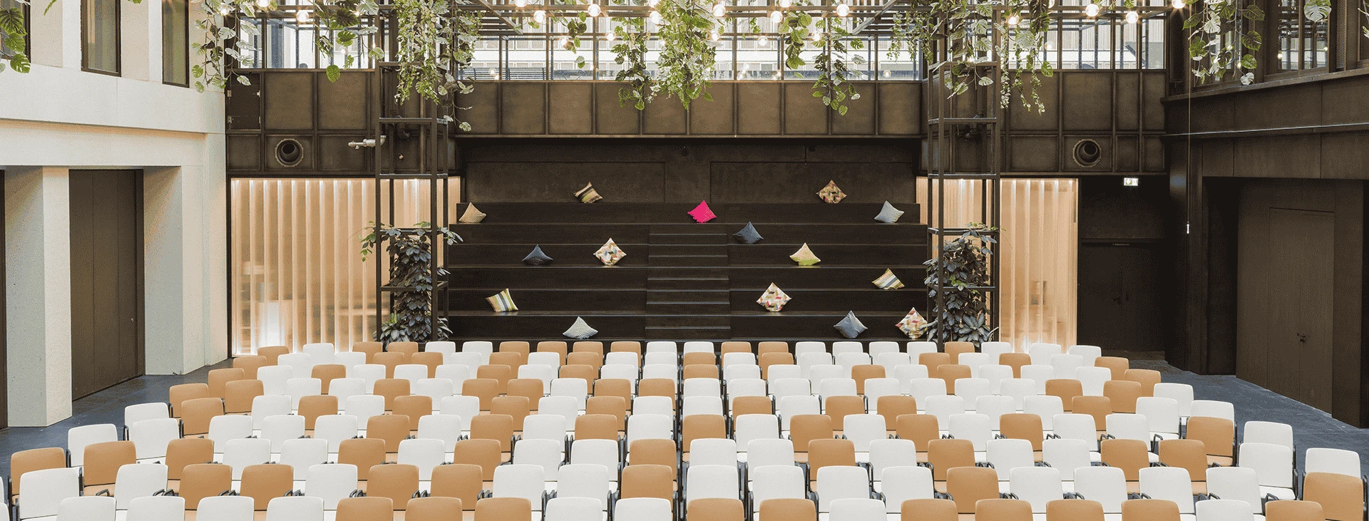 Modern eingerichteter Veranstaltungsraum mit Reihen von Stühlen, gerichtet auf eine Bühne, umgeben von hängenden Pflanzen und Lichterketten an der Decke.