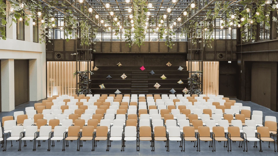 Modernly furnished event hall with rows of chairs facing a stage, surrounded by hanging plants and string lights on the ceiling.