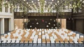 Modernly furnished event hall with rows of chairs facing a stage, surrounded by hanging plants and string lights on the ceiling.