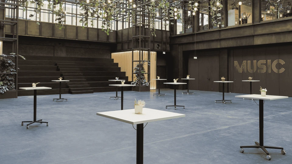 Interior view of a modern event venue with numerous empty tables set on a blue carpeted floor. In the background, a large staircase and the illuminated word 'MUSIC' on the wall.