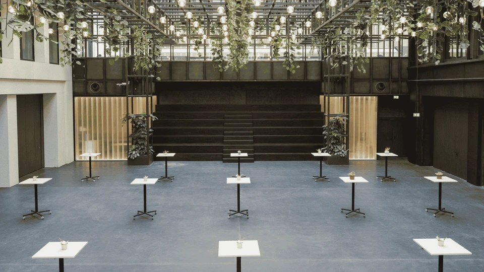 Modern interior of a building featuring a high glass ceiling, numerous plants, several tables, and tiered seating in the background.