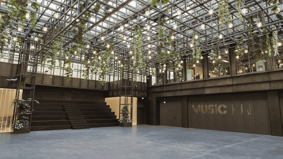 Modern hall with a glass roof through which plants hang, illuminated by numerous bulbs, featuring a large staircase and the inscription 'WERK STATT' on a wall.