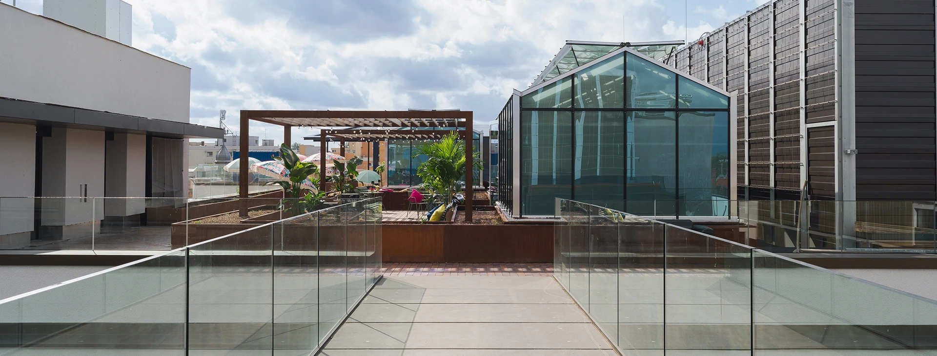 Modern rooftop terrace with a glass building, surrounded by other structures, plants, and a pergola under a cloudy sky.
