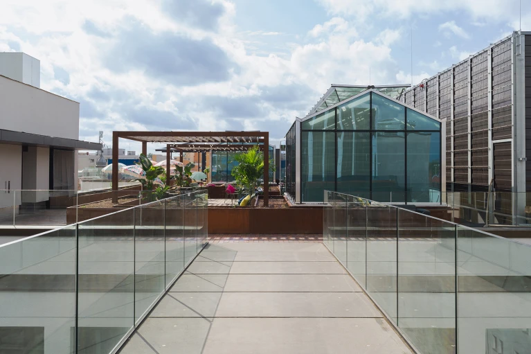 Moderne Dachterrasse mit Glasgebäude, umgeben von anderen Gebäuden, Pflanzen und einer Pergola unter einem bewölkten Himmel.