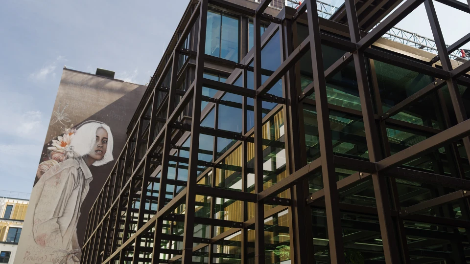 Ansicht der Außenfassade mit moderner Stahl-Glas-Konstruktion, im Hintergrund ein großformatiges Wandgemälde mit Frau und Blumen.
