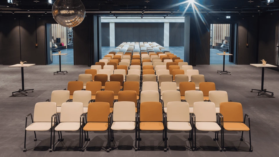 Moderne Konferenzsaal mit Reihen von orangen und beigen Stühlen gerichtet auf eine Bühne im Hintergrund, beleuchtet von Deckenspots.