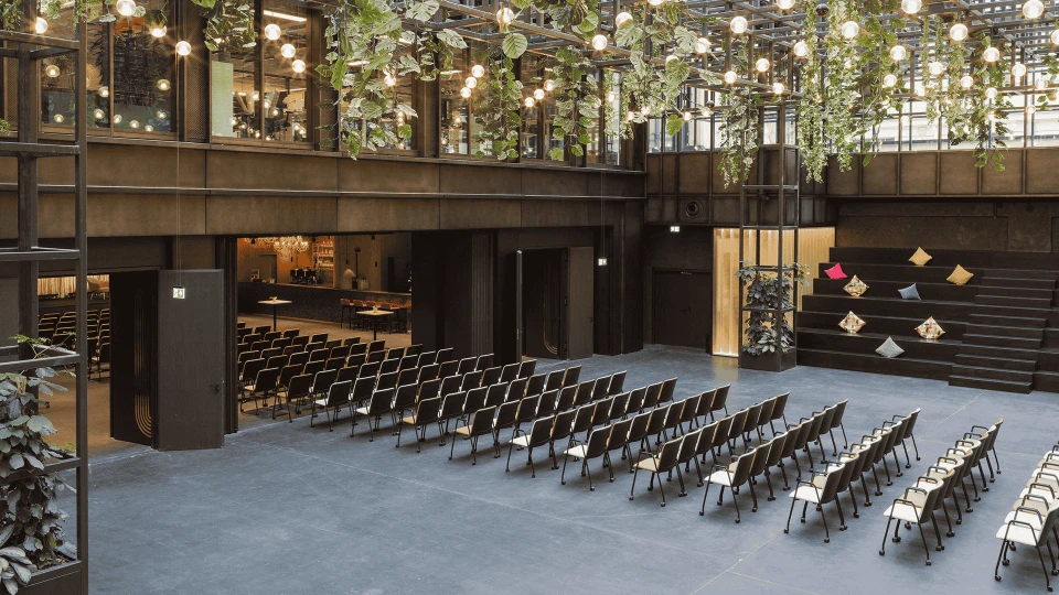 Moderne Veranstaltungshalle mit Reihen schwarzer Stühle, angeordnet für ein Publikum, umgeben von einer metallischen Struktur mit hängenden Pflanzen und Leuchten
