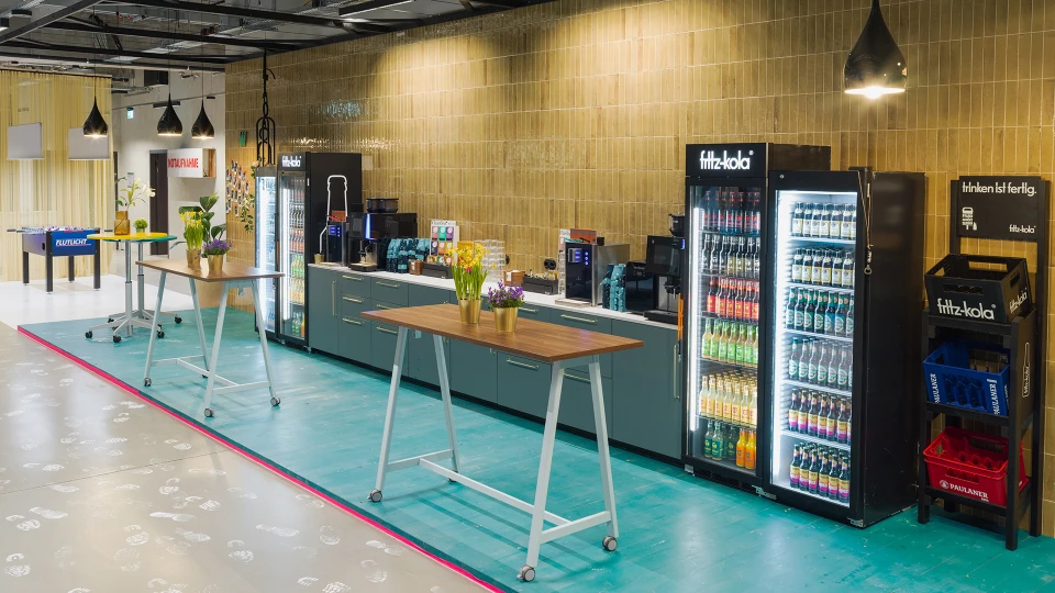 Getränkestation mit Kühlschränken von Fritz-Kola, Kaffeemaschinen, bunten Tischen mit Blumen und hängenden Lampen vor grün gefliester Wand.