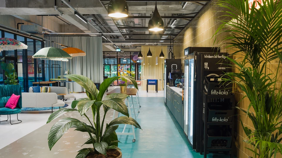 Weitwinkelaufnahme eines farbenfrohen Lounge- und Barbereichs mit großen Pflanzen, Theke mit Getränken, Sitzgelegenheiten und modernen Deckenleuchten.