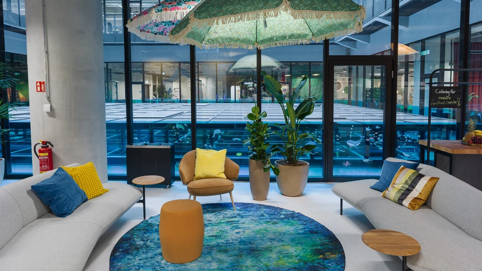 View of a creatively designed lounge area featuring a glass cabin, colorful umbrellas, sofas, plants, and a view of the open architecture.