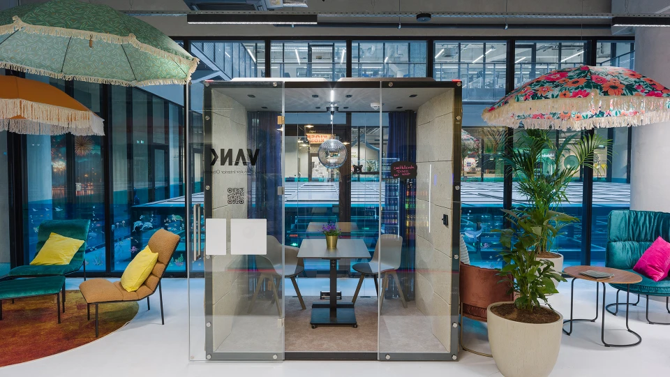 Glaskabine mit Diskokugel und kleinem Tisch mit vier Stühlen, umgeben von bunten Sonnenschirmen, Sesseln und Pflanzen im Lounge-Stil.