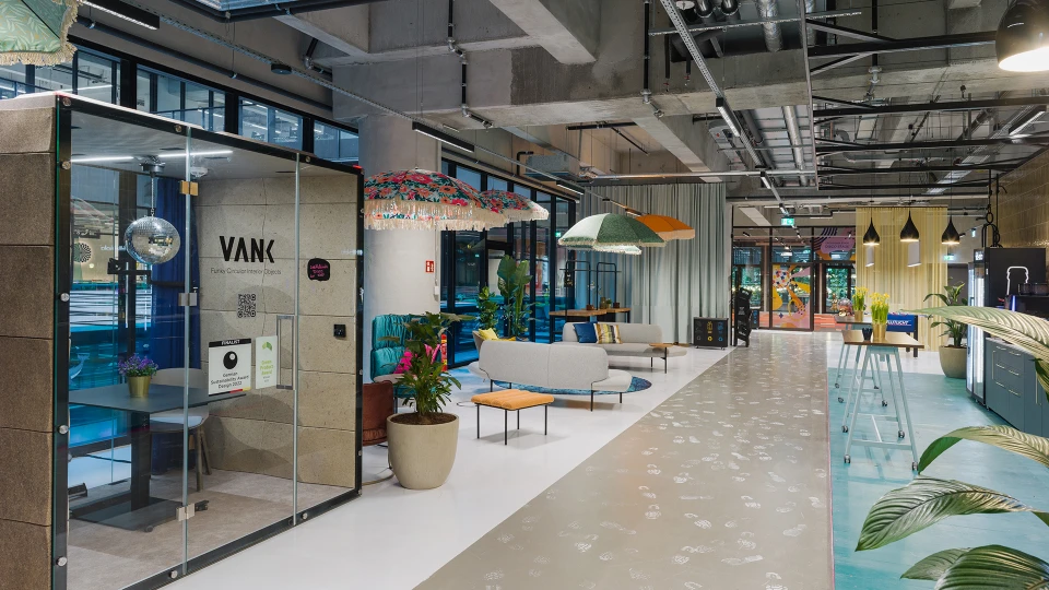 Weitläufige Ansicht eines kreativ gestalteten Loungebereichs mit Glaskabine, bunten Schirmen, Sofas und Pflanzen sowie einem Blick auf die offene Architektur.