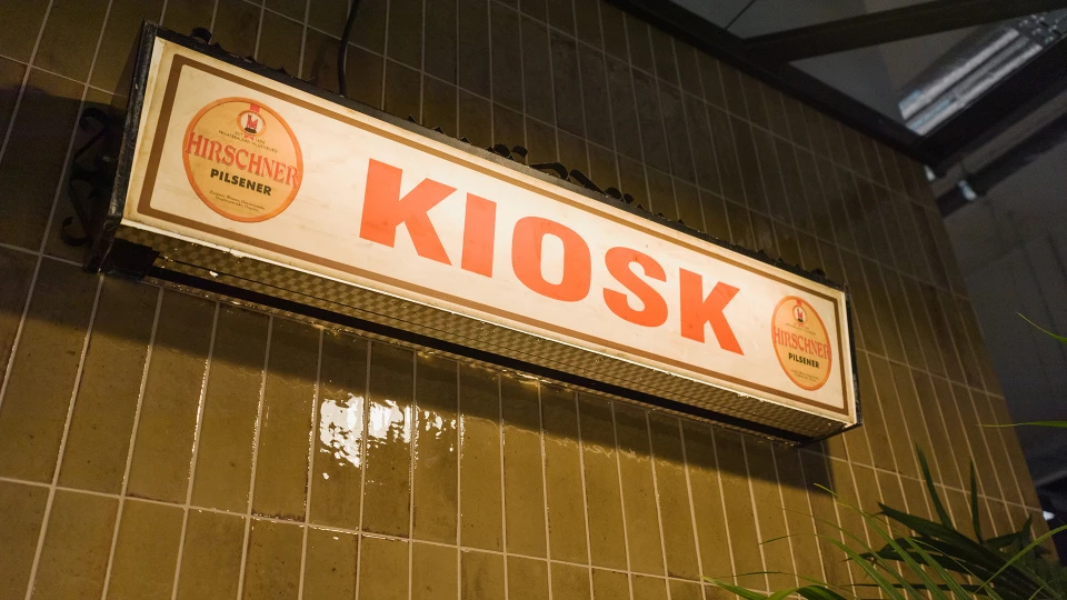 Vintage-Leuchtschild mit der Aufschrift „KIOSK“ und Hirschner Pilsener Logos, montiert an einer Wand mit grünen Keramikfliesen.