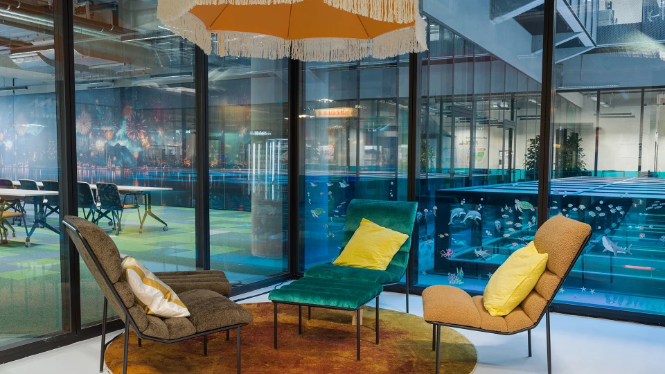 Kleiner, gemütlicher Sitzbereich mit Sesseln unter einem orangefarbenen Sonnenschirm, Blick durch Glaswände auf ein Raum mit Feuerwerksmotiv an der Wand.