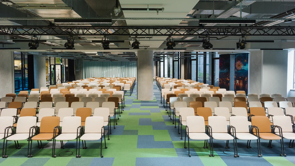 Blick in einen großen Konferenzraum mit Reihenbestuhlung aus beigen und weißen Stühlen auf grün-blau gemustertem Teppichboden.