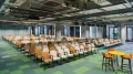 Konferenzraum mit gestaffelter Reihenbestuhlung in hellen Farben; im Vordergrund ein gelber Stehtisch mit zwei orangefarbenen Hockern.