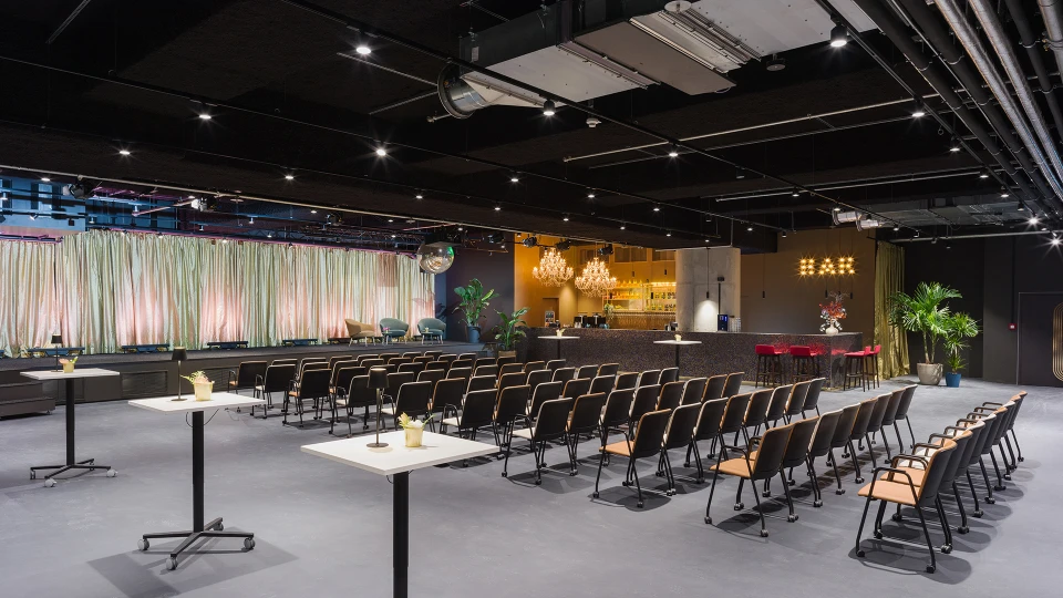 Moderne Veranstaltungshalle mit Reihen von schwarzen Stühlen, einer beleuchteten Bar im Hintergrund und dekorativen Elementen
