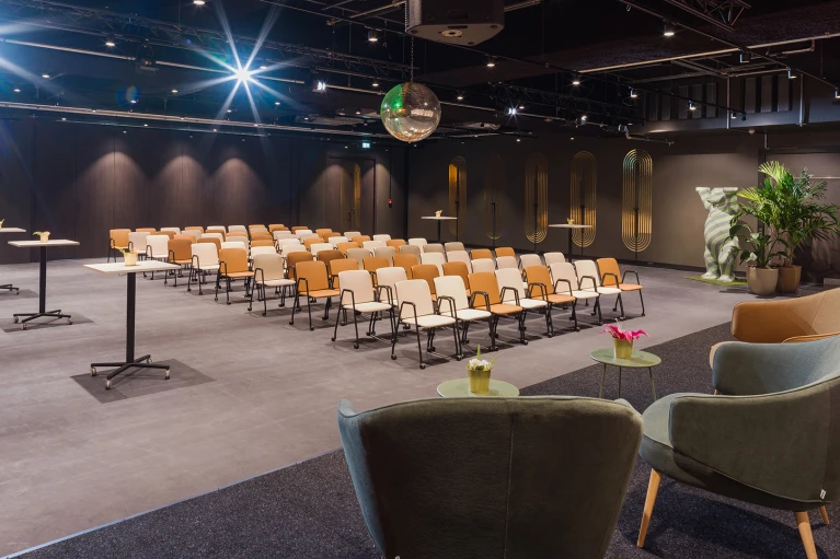 Modern gestalteter Konferenzraum mit Reihen von beige-braunen Stühlen, Stehtischen, Grünpflanzen und einer Diskokugel an der Decke