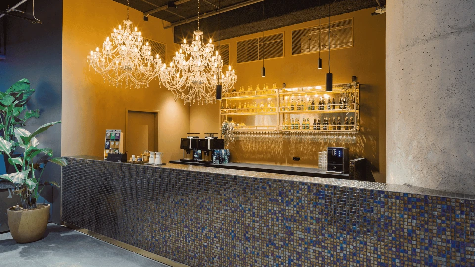 Eleganter Innenraum einer Bar mit Mosaik-Tresen, darüber zwei große Kronleuchter und Regale voller Gläser und Flaschen.