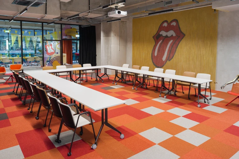 Meetingraum mit U-förmig gestellten Tischen, orangerotem Teppich, markanter Zungen-Grafik an der Wand und Sitzfigur mit Schallplatte.
