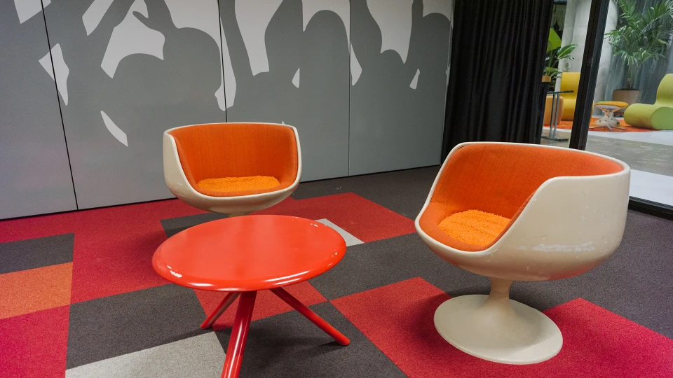 Zwei retrofuturistische Lounge-Stühle mit oranger Polsterung und kleinem roten Tisch auf einem rot gemusterten Teppich.