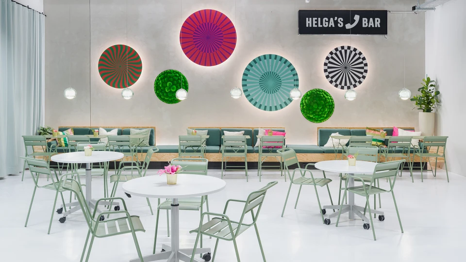 Modernly designed bar with green and white furniture, decorated with colorful wall fans and labeled 'Helga's Bar'.