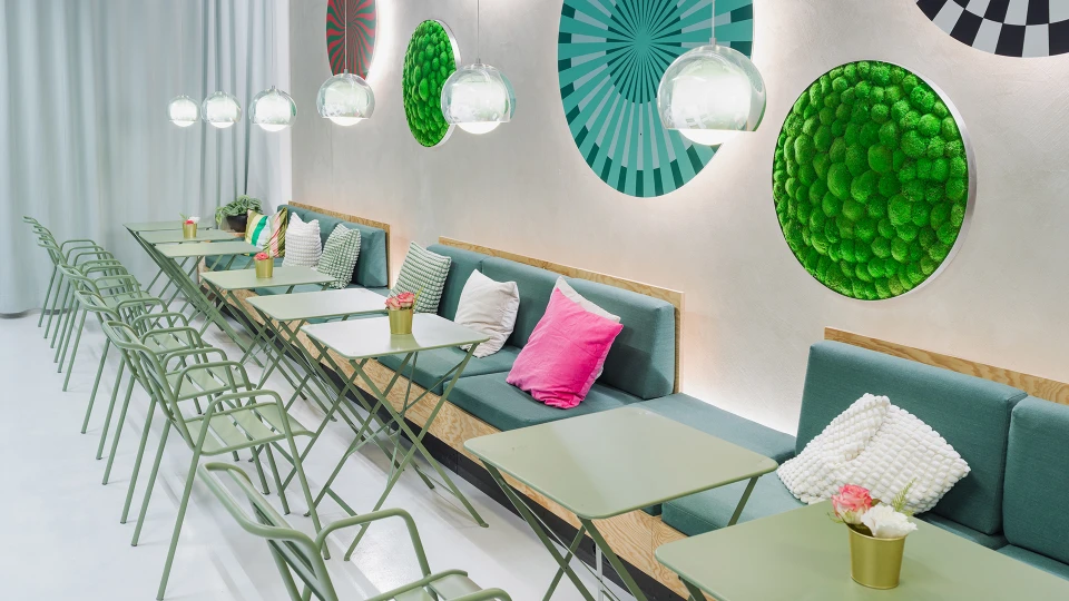View of a cozy seating area with green folding chairs and light green tables in front of a wall adorned with circular graphic artworks and moss installations.