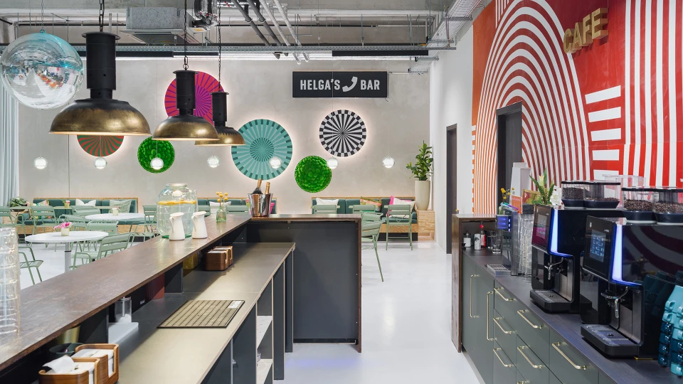 Interior of a cafe with a bar counter in the foreground, and a background featuring a mix of modern seating groups, circular wall installations, and a 'Helga's Bar' sign.