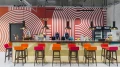 Central front view of a bar with a gold-colored counter, colorful bar stools, and a red-white wall design with the word 'Café' above the door.