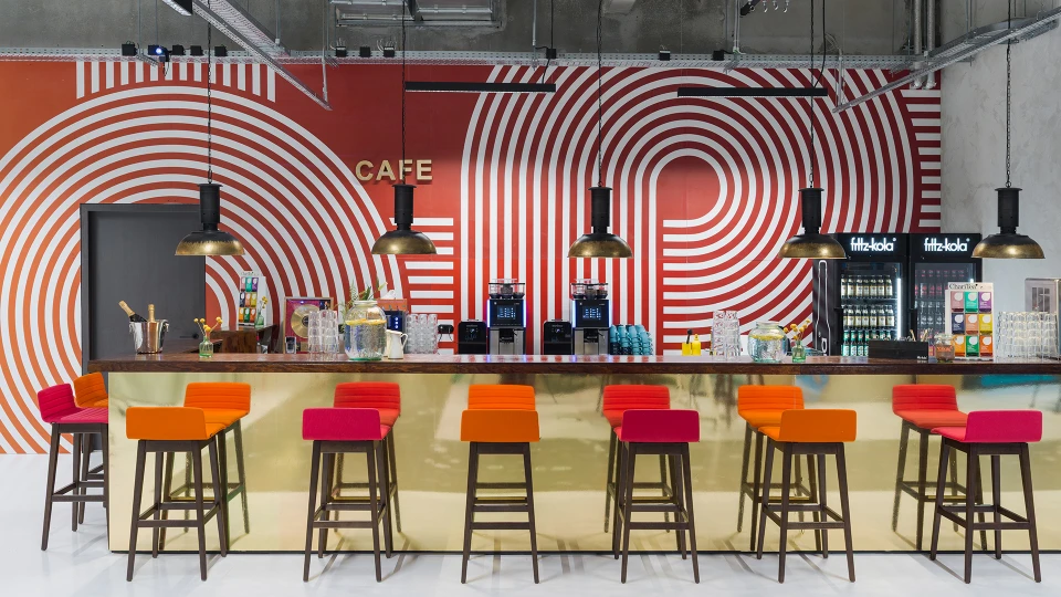 Central front view of a bar with a gold-colored counter, colorful bar stools, and a red-white wall design with the word 'Café' above the door.