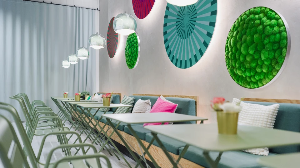 Detailansicht der langen Sitzbank mit mintfarbenen Kissen, runden Wandinstallationen und hängenden Glaskugelleuchten in einem hellen Cafébereich.