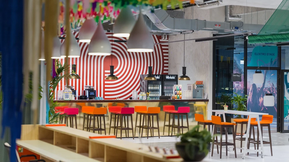 Colorful overall view into the café through a decoration, focusing on the bar area with colorful stools, lighting fixtures, and beverage coolers.