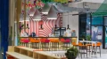 Colorful overall view into the café through a decoration, focusing on the bar area with colorful stools, lighting fixtures, and beverage coolers.