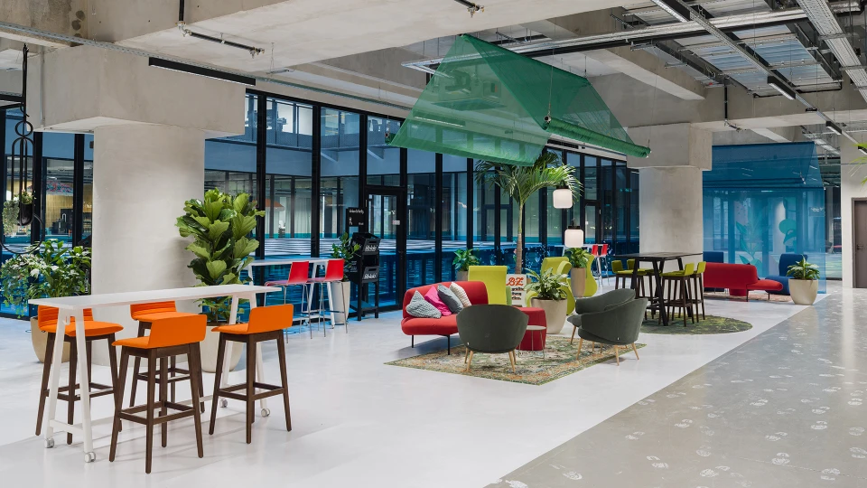 Lounge-Bereich mit roten und blauen Sofas, Teppich, Pflanzen und einem blauen Netzdach unter hoher Glasfassade.