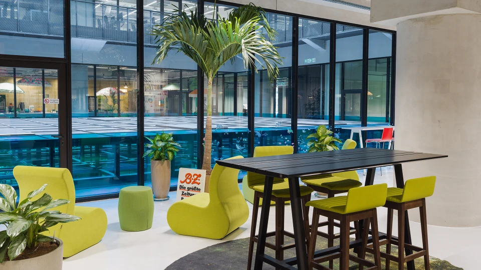 Modern eingerichtete Lounge mit hohen Barstühlen an einem schwarzen Tisch, grünen Sitzsäcken und großen Topfpflanzen vor einer Glasfront mit Blick auf einen Poolbereich